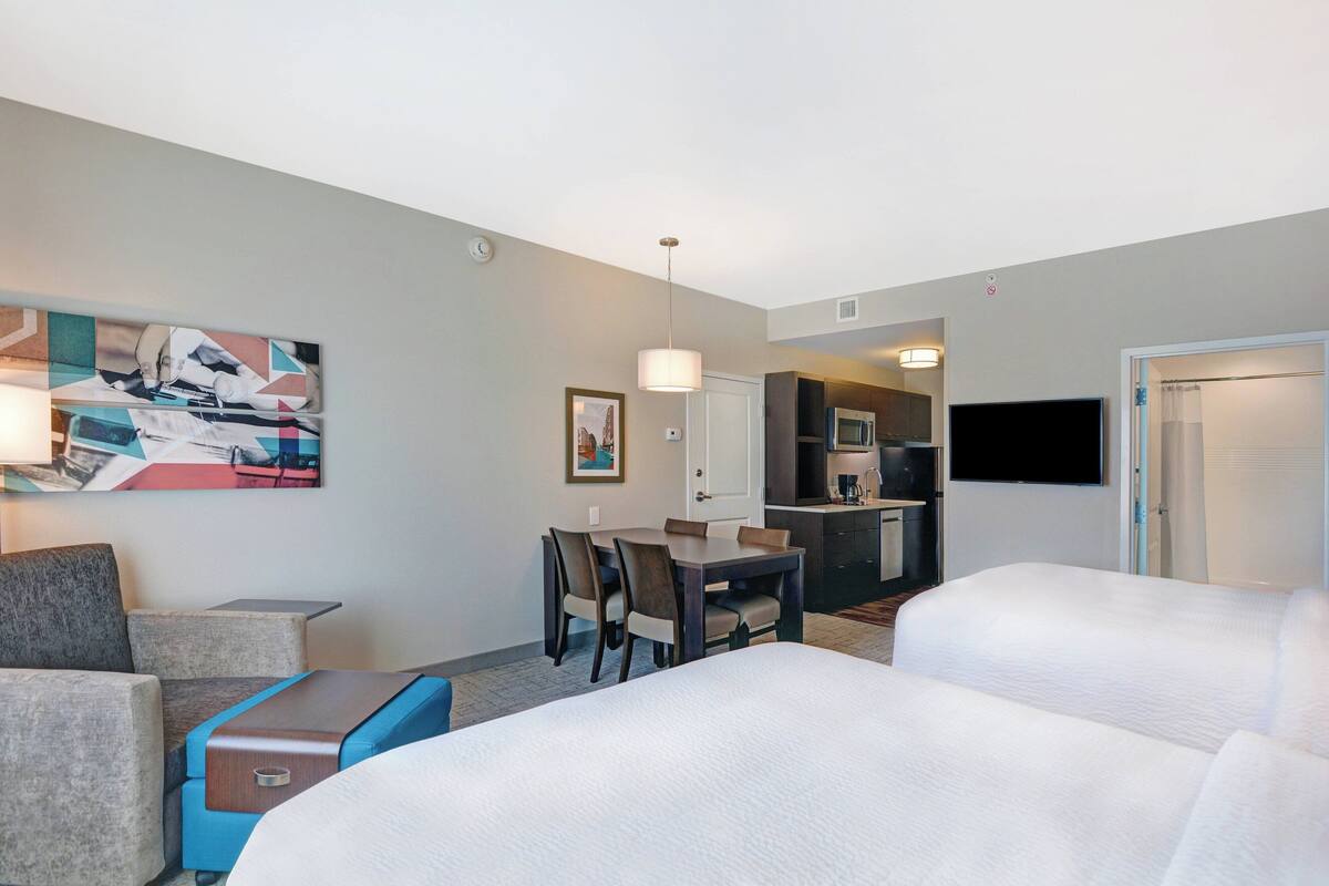 Una habitación de hotel con dos camas, una zona de comedor con mesa y sillas, una kitchenette y un televisor en la pared.