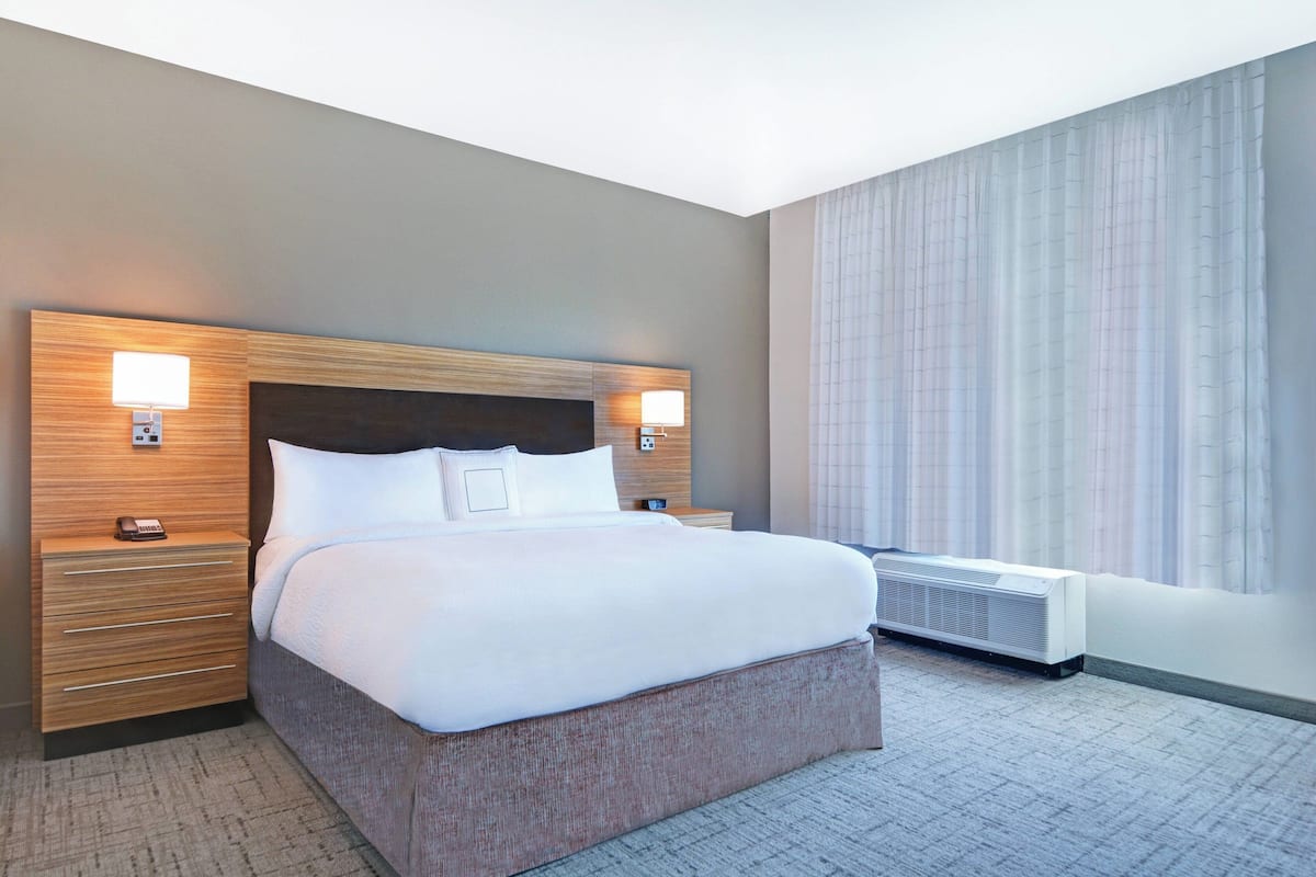 Una habitación de hotel con una cama grande, dos mesitas de noche con lámparas, una ventana con cortinas y un equipo de aire acondicionado.