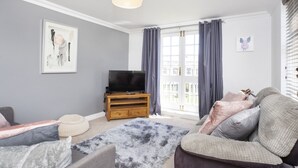 A living room with a flat-screen TV on a wooden stand, a grey sofa, a patterned rug, and a large window with curtains. - Fountains View Apartment (York)