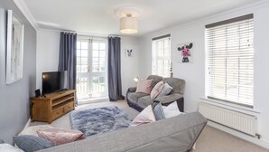 A well-lit room with a sofa, a television on a wooden stand, and a window with blinds. - Fountains View Apartment (York)