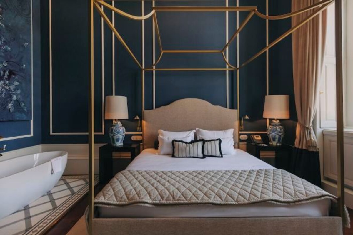 A bedroom with a four-poster bed, bedside tables with lamps, a large window with curtains, and a wall-mounted artwork.