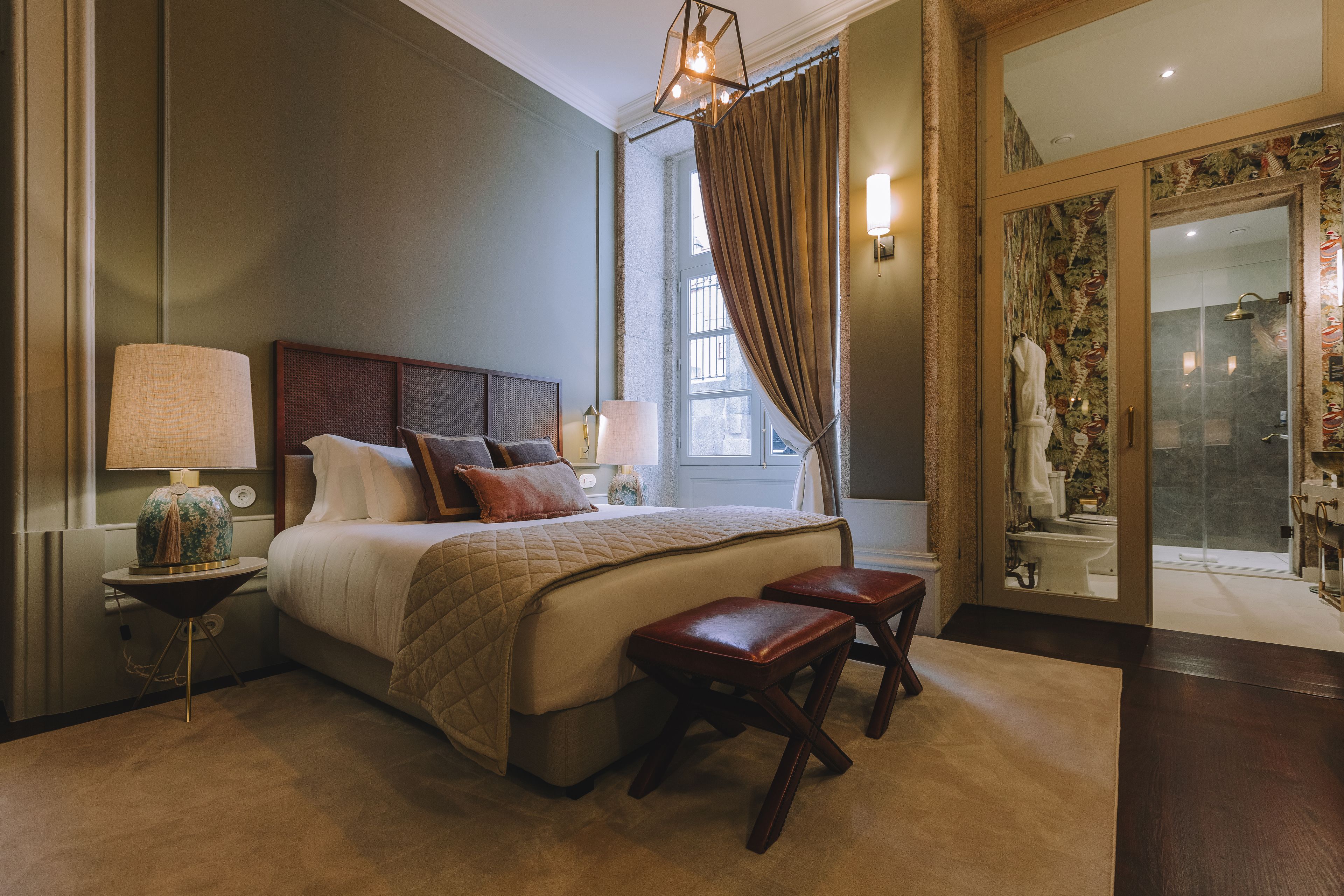 A hotel room with a large bed, a bedside table with a lamp, a small stool, and a view of the bathroom through a glass door.