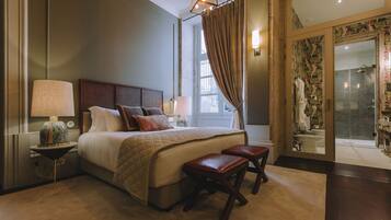 A hotel room with a large bed, a bedside table with a lamp, a small stool, and a view of the bathroom through a glass door.