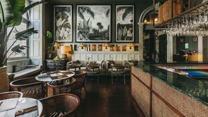 Interior de restaurante con mesas altas, sillas de mimbre y una zona de bar con vasos colgantes.