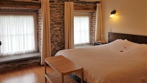 Een slaapkamer met een stenen muur, houten plafond, een bed met een witte deken, een houten bankje en een nachtkastje met een lamp.