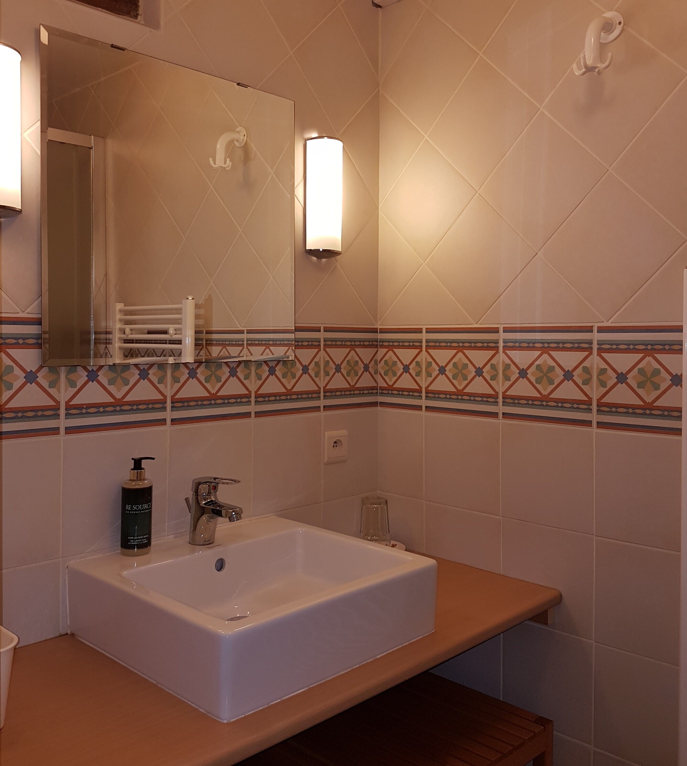 Een badkamer met een rechthoekig wastafelblad, een spiegel, een wandlamp en een decoratieve tegelrand.