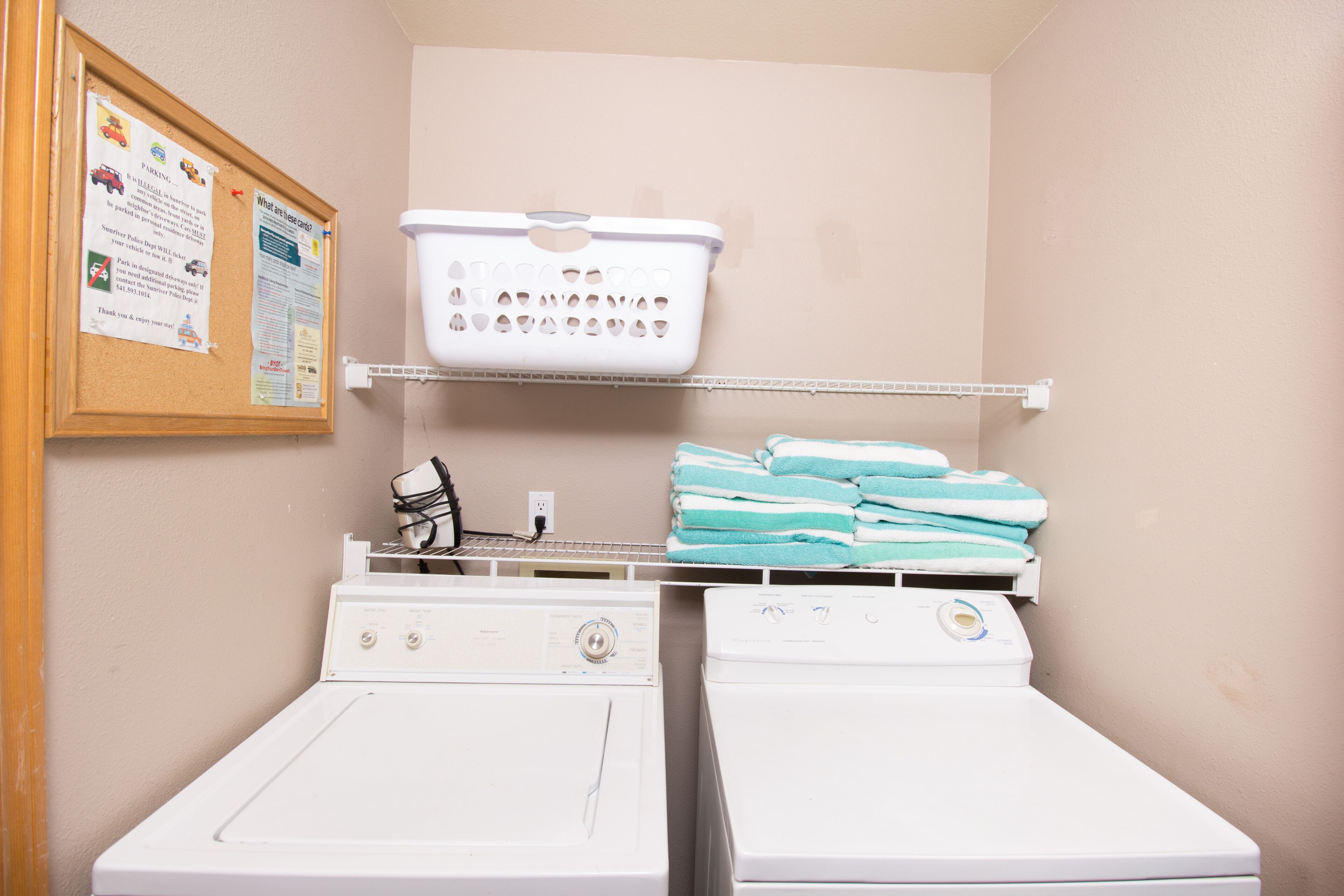 Une buanderie équipée de deux machines à laver et deux sèche-linge blancs, d’un tableau d’affichage où sont accrochées des annonces, et d’une étagère où sont pliées des serviettes.