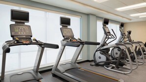 Eine Reihe von Laufbändern mit digitalen Bildschirmen und Bedienelementen, aufgestellt in einem Fitnessstudio mit großen Fenstern und einer Decke mit Leuchtstofflampen.