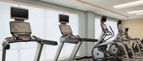 Une rangée de tapis de course dotés d’écrans numériques et de panneaux de commande, disposés dans une salle de sport avec de grandes fenêtres et un plafond équipé de néons.