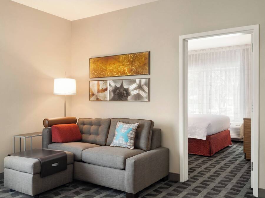 Habitació d'hotel moderna amb un sofà seccional gris, una taula petita, una làmpada i obra d'art a la paret.