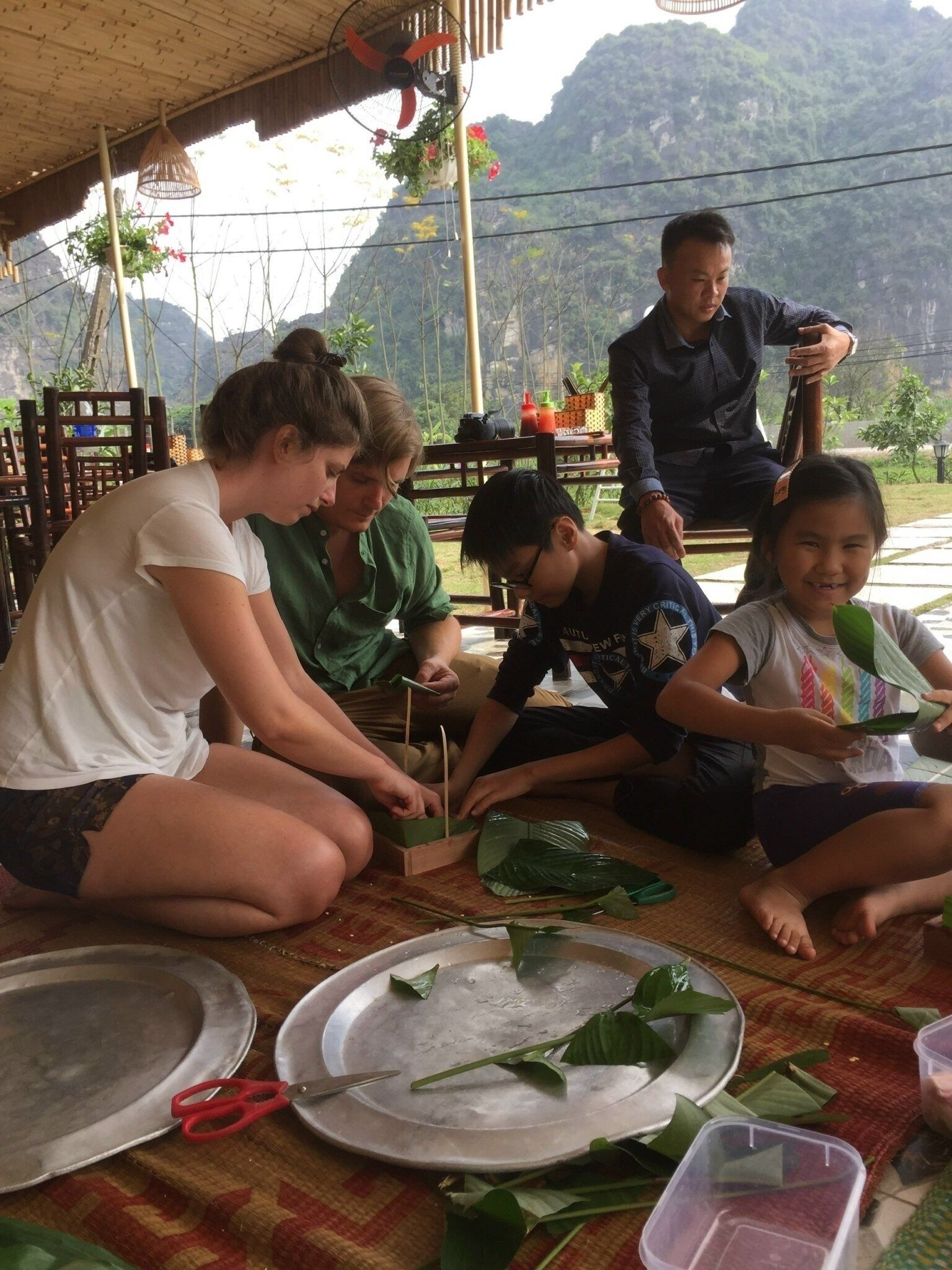 A group of people engaged in an outdoor activity, possibly a craft or cooking session, with natural scenery in the background.
