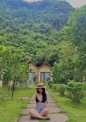 A person sitting on a stone pathway in a lush garden with a house in the background. - Trang An Retreat (Tay Hoa Lu)
