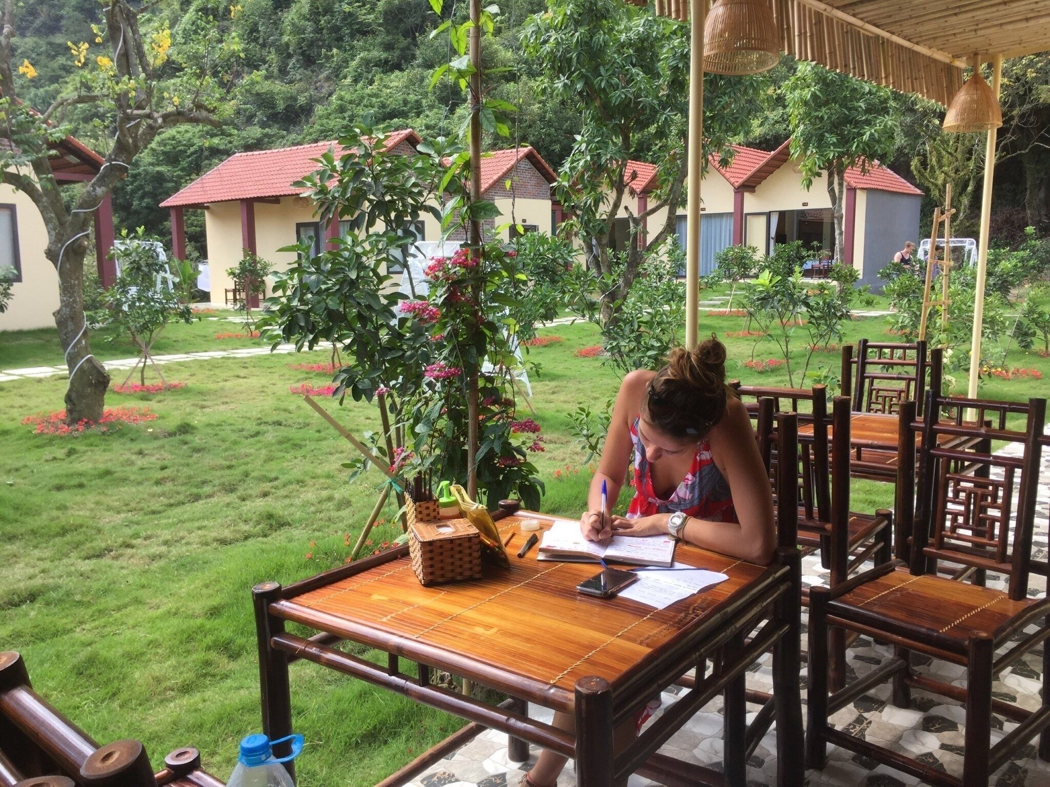 Une femme est assise à une table en bois à l’extérieur, avec un carnet à portée de main, entourée de chaises et d’une plante en pot.