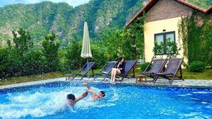 A swimming pool with two people swimming, surrounded by lounge chairs and a building with a tiled roof. - Trang An Retreat (Tay Hoa Lu)