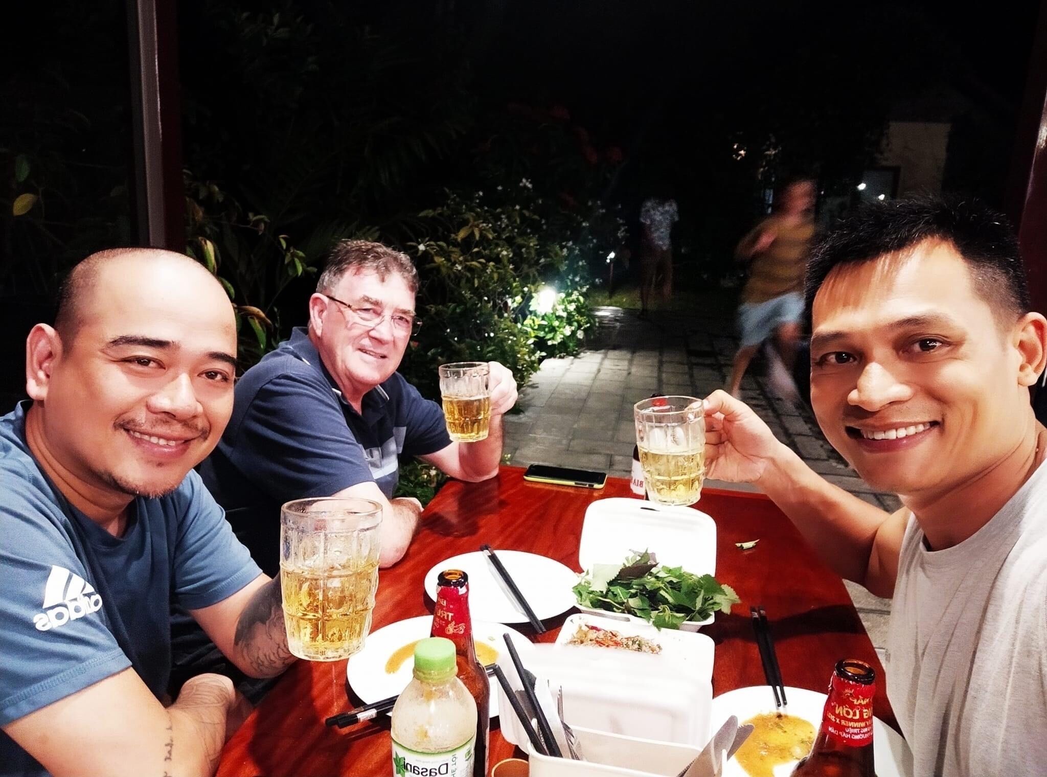 Trois personnes sont assises à une table, avec des boissons et de la nourriture, à l’extérieur, la nuit.