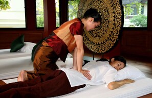 A person receiving a massage in a room with a large decorative wall hanging, a bed with white bedding, and a window with a view of greenery.