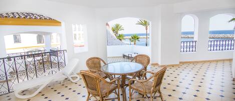 Una habitación iluminada por el sol, con piso de baldosas, una mesa redonda con sillas y vistas al mar a través de ventanas arqueadas.