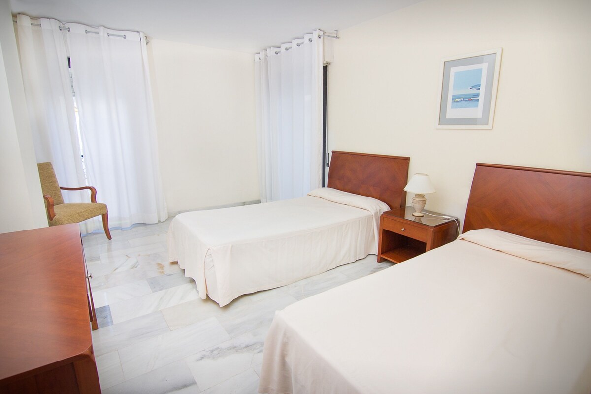 Een hotelkamer met twee enkele bedden, een houten nachtkastje met een lamp, een stoel en een ingelijst kunstwerk aan de muur.