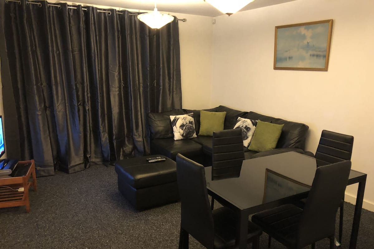 Un salon avec un canapé en cuir noir, des coussins verts, un pouf noir et une table à manger en verre avec des chaises noires.