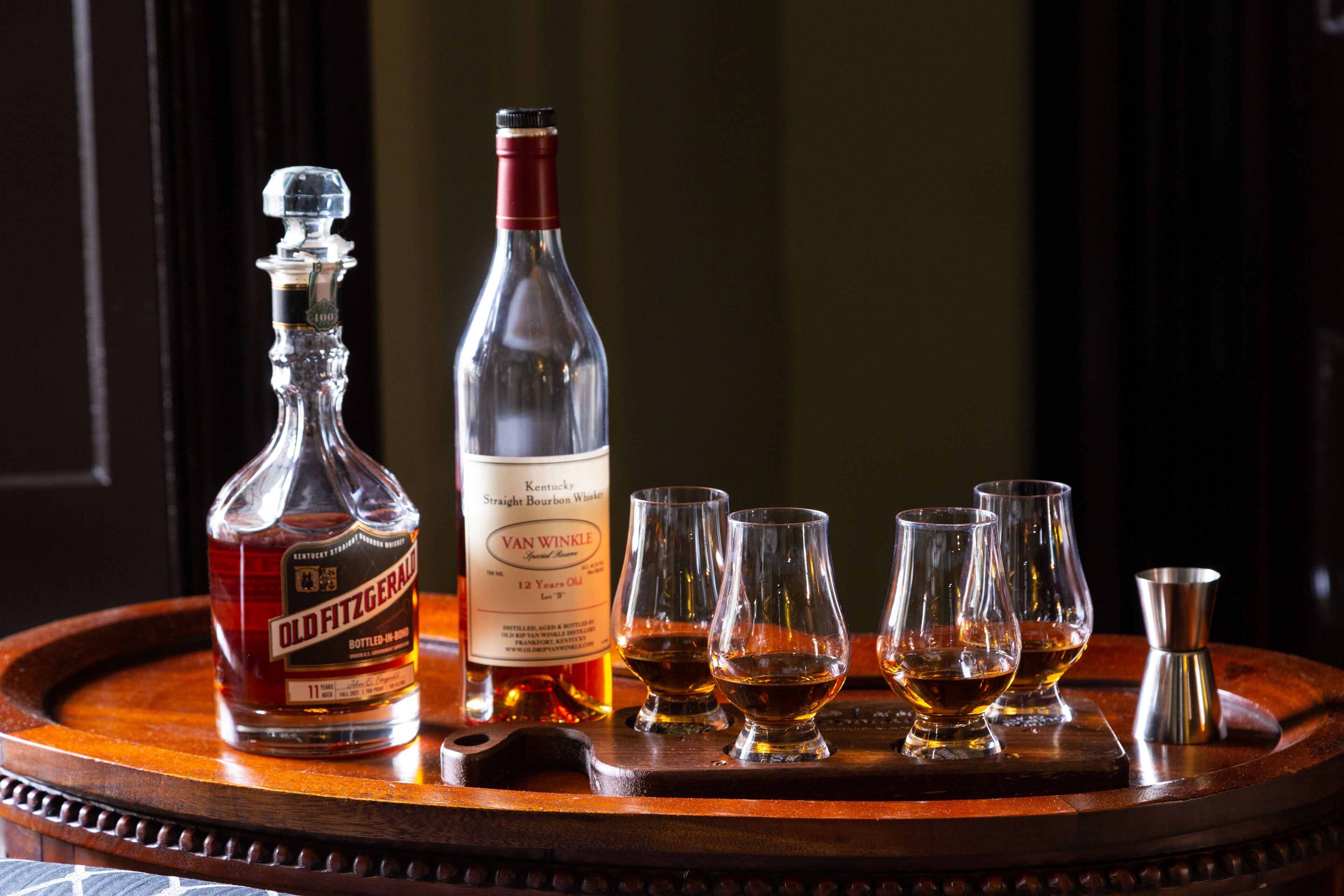 A wooden tray with a bottle of Old Fitzgerald Bourbon Whiskey, a bottle of Van Winkle, and four glasses of whiskey.
