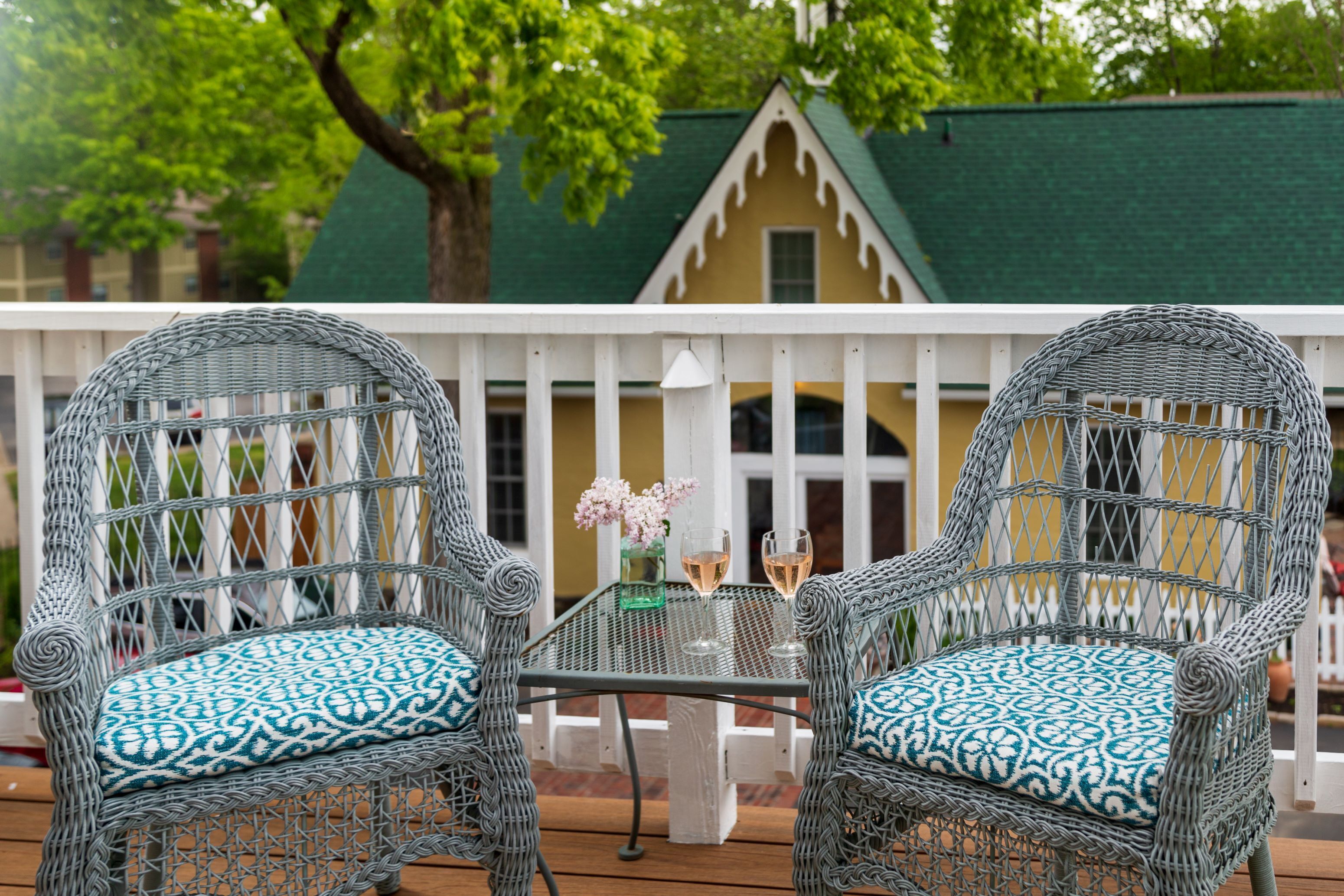 Two wicker chairs with patterned cushions on a wooden deck, a table with two glasses of wine, and a building with a green roof in the background.