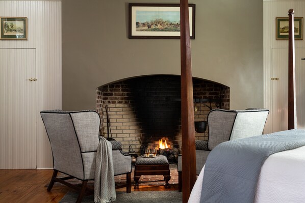 A room with a brick fireplace, two upholstered chairs, a wooden four-poster bed, and a framed picture on the wall.