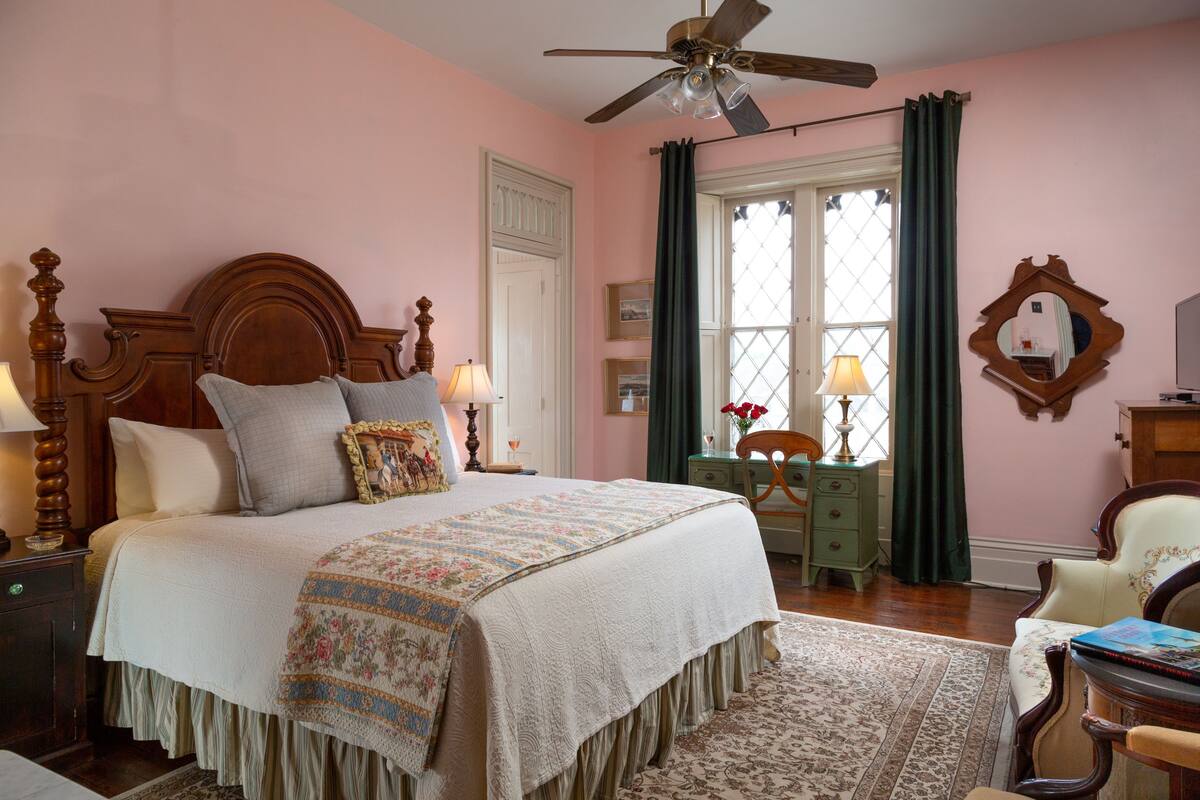 Une chambre avec un grand lit, un ventilateur de plafond, une fenêtre avec des rideaux, un bureau avec une lampe et une chaise.