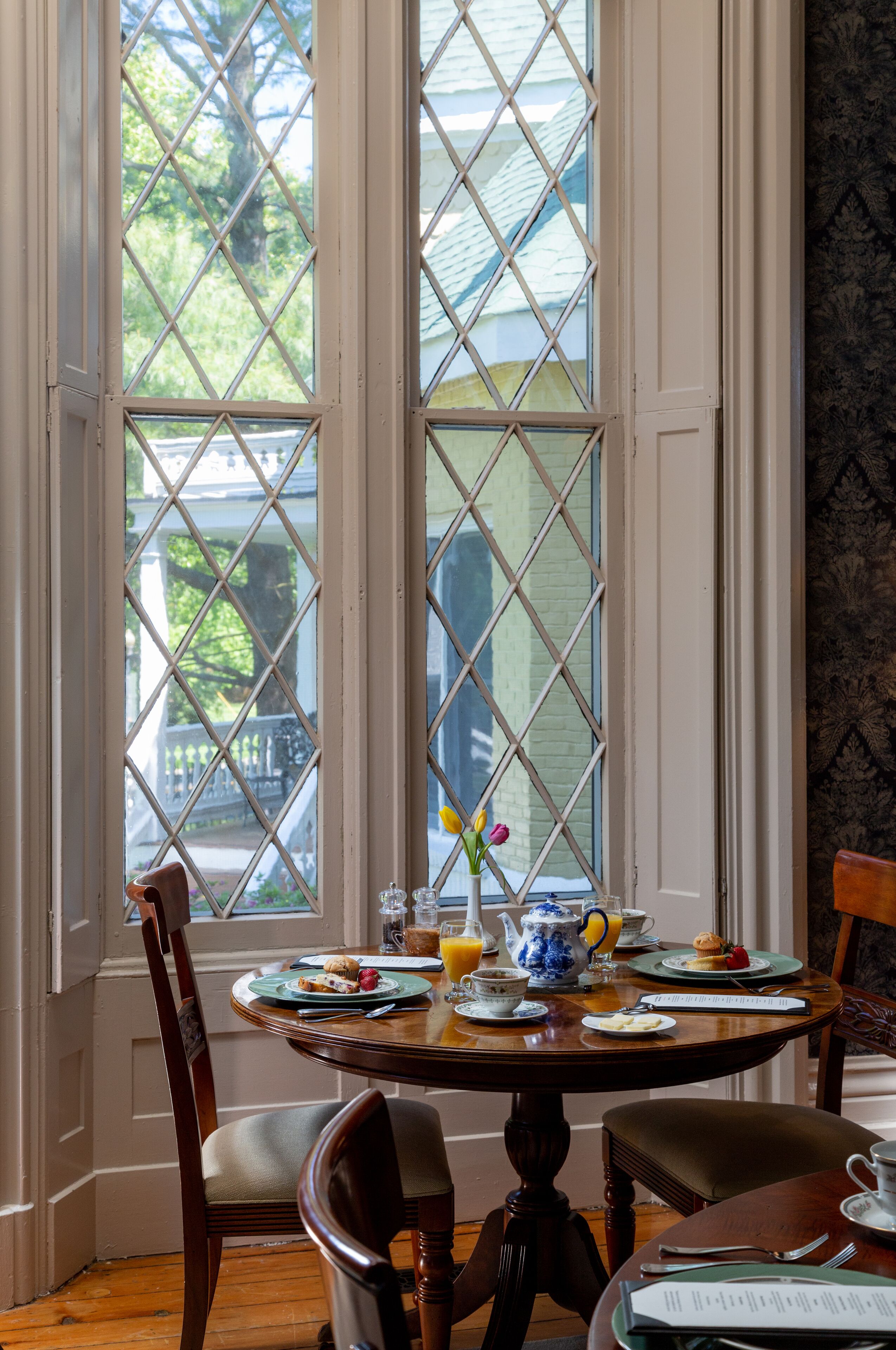 A dining area with a round table set for breakfast, a window with a view of greenery, and a patterned wall.