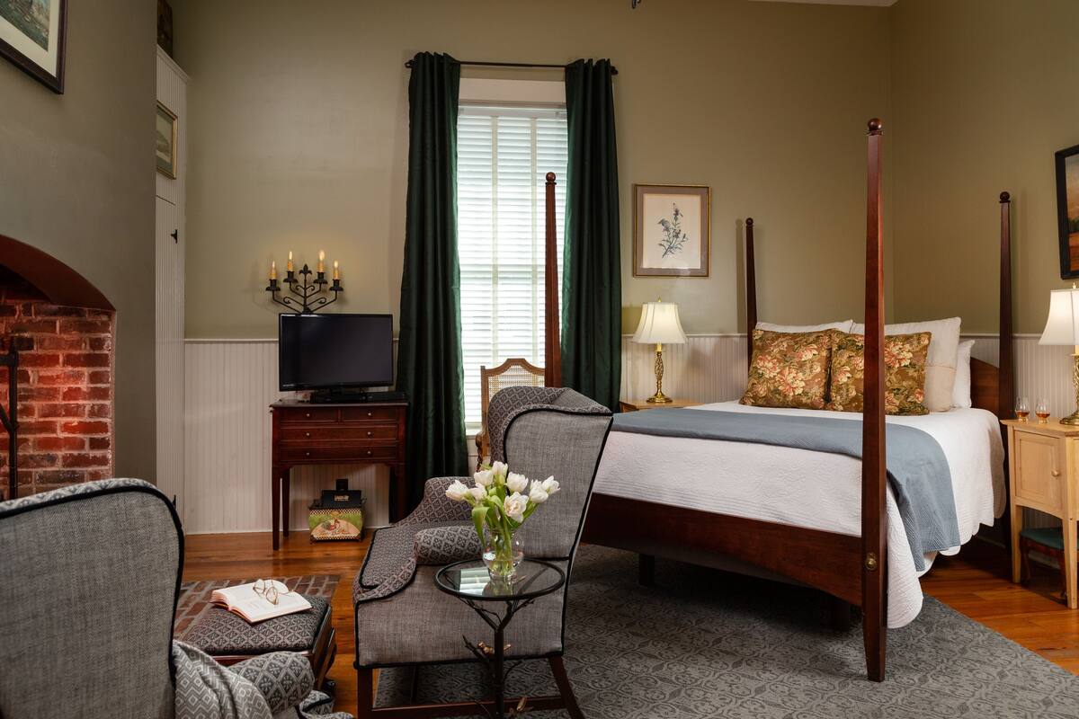 Une chambre à coucher avec un lit à baldaquin, un téléviseur posé sur un meuble en bois, une chaise agrémentée d’un vase de fleurs et une cheminée.