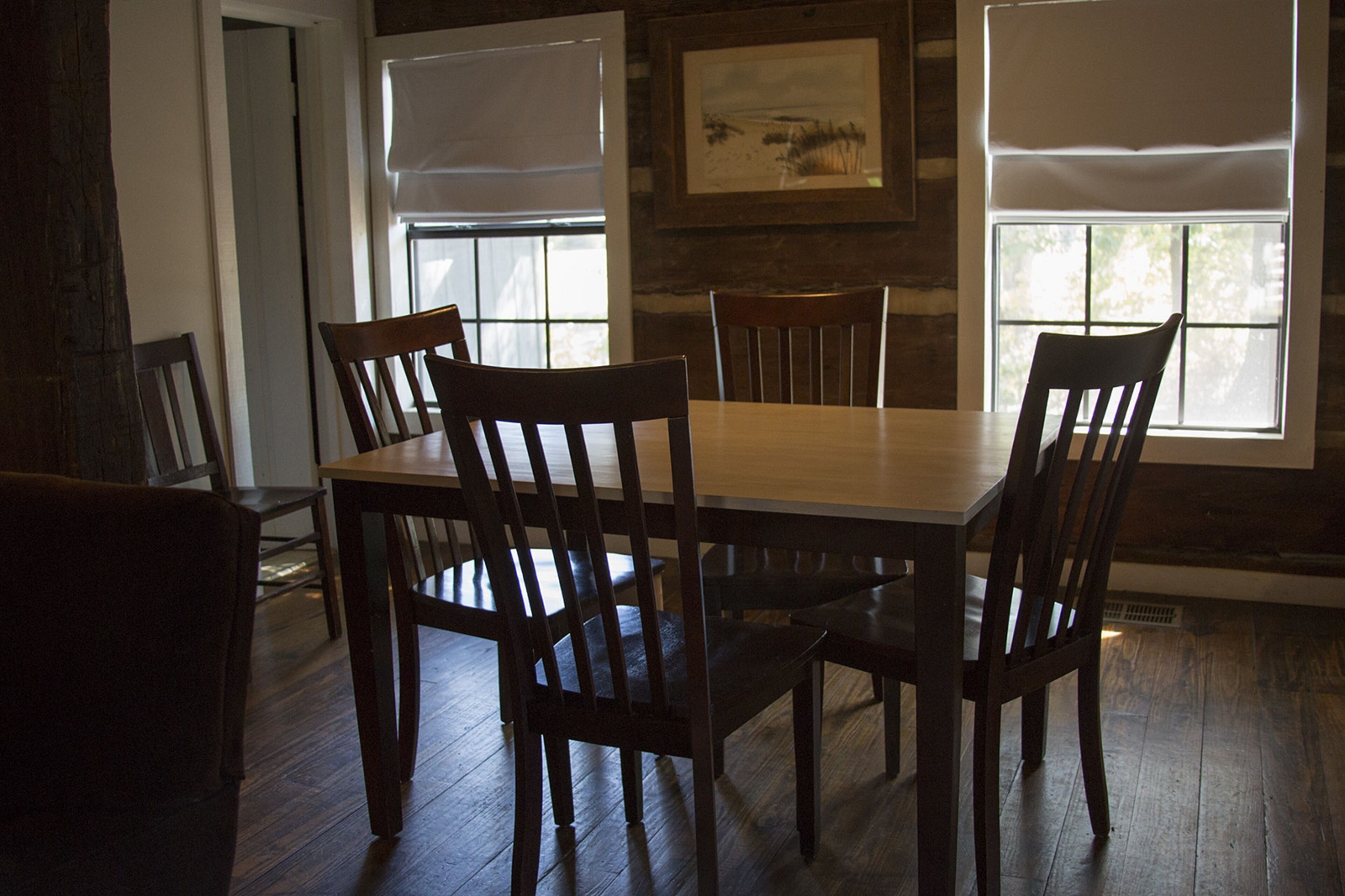 A dining area with a wooden table and chairs, large windows with blinds, and a framed picture on the wall.