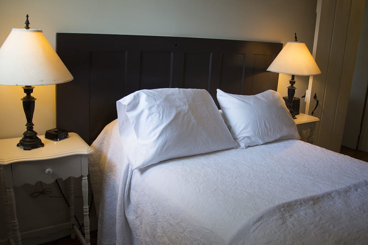 A neatly made bed with white linens and pillows, flanked by two bedside tables with lamps, in a room with a dark headboard.