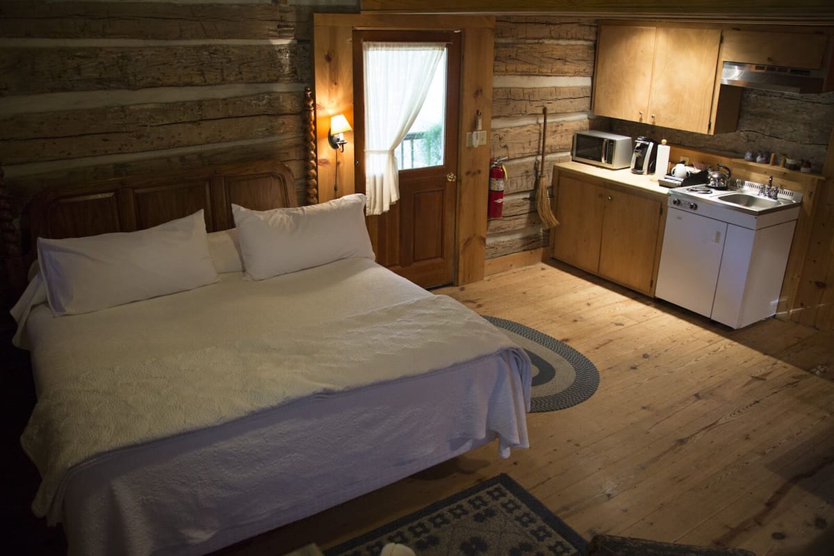 A cozy log cabin bedroom with a large bed, wooden walls, a small kitchenette, and a door leading to a balcony.
