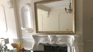 A dining area with a wooden table, chairs, a vase with flowers, a white fireplace with a gold-framed mirror above it, and a chandelier. - The Rhinos 2 (Enfield)