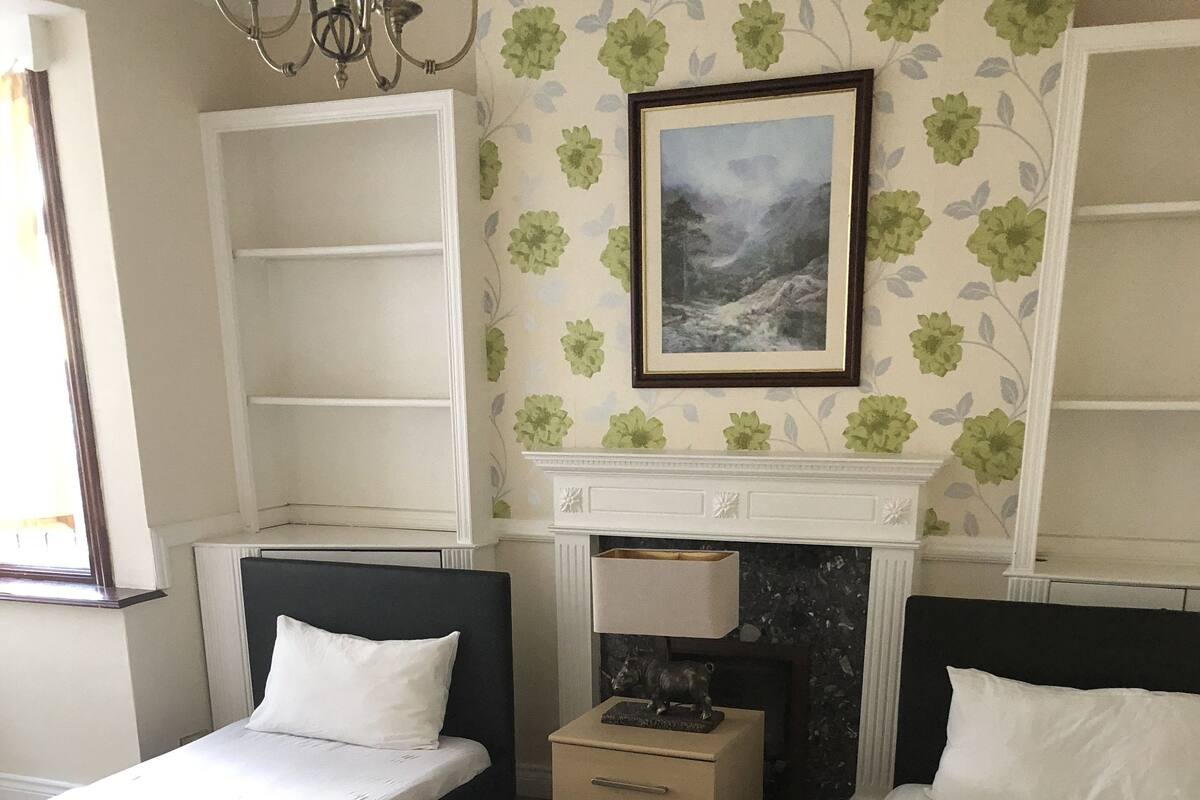 A bedroom with two single beds, a nightstand with a lamp, a fireplace, and floral wallpaper.