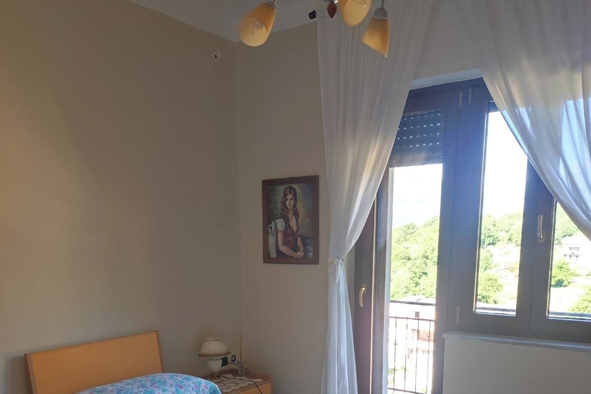 A bedroom with two beds, a nightstand, a lamp, a painting on the wall, and a balcony with curtains.