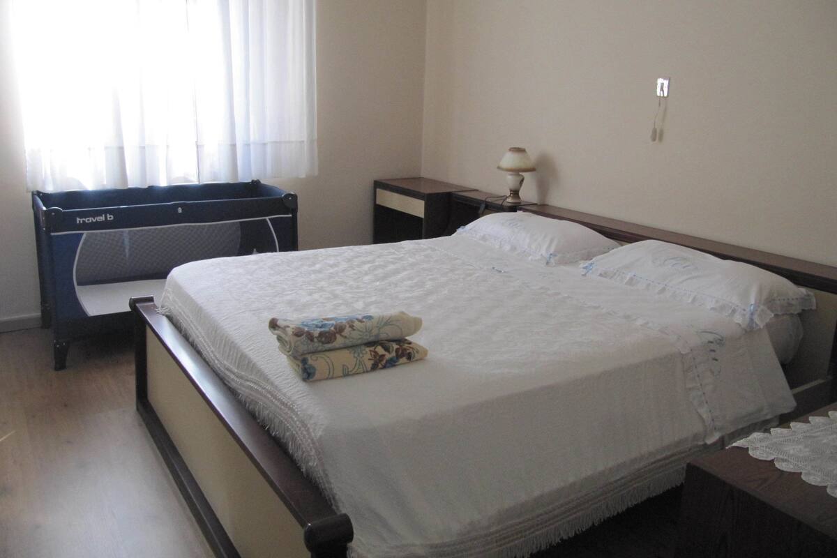 A bedroom with a large bed, a bedside table, a lamp, and a window with curtains.