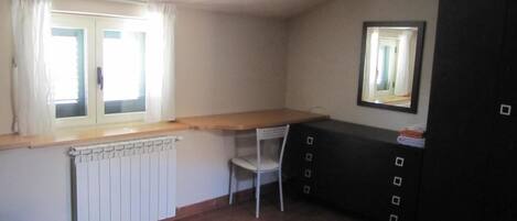 A small room with a radiator, a window with curtains, a chair, a desk with a mirror, and a cabinet.