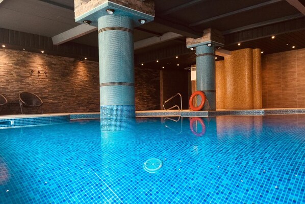 A swimming pool with blue mosaic tiles, supported by blue pillars, and surrounded by wooden walls and a brick wall. - THE Cumberland (Bournemouth)
