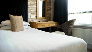 Habitación de hotel con cabecera de madera, cama con ropa blanca, cómoda de madera, una silla, un espejo y ventana con vistas.