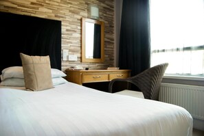 A hotel room with a wooden headboard, a bed with white linens, a wooden dresser, a chair, a mirror, and a window with a view. - THE Cumberland (Bournemouth)