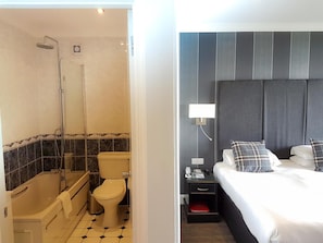 A bathroom with a bathtub, toilet, and shower, and a bedroom with a bed, nightstand, and wall-mounted lamp. - THE Cumberland (Bournemouth)