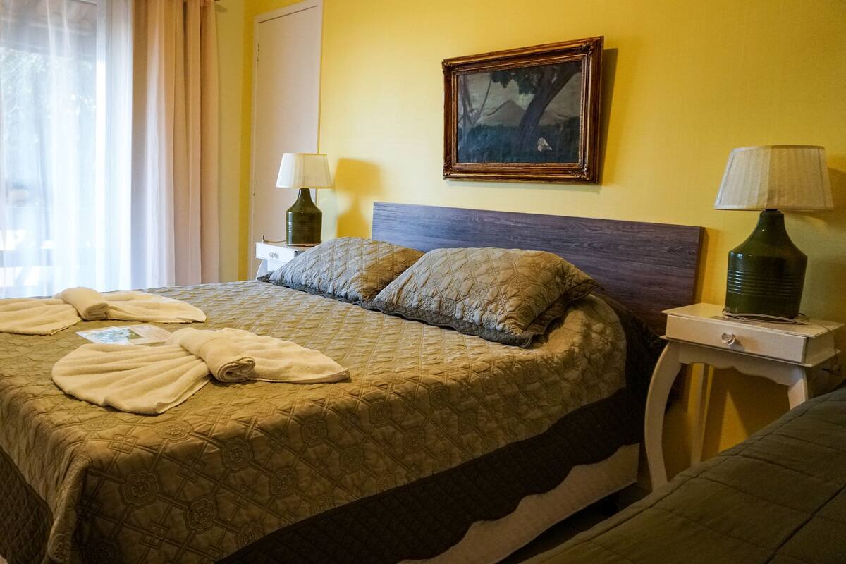 Un lit bien fait, avec une couvre-lit matelassé, deux oreillers et des serviettes enroulées. Il y a un tableau accroché au mur au-dessus du lit, une lampe sur une table de chevet et une fenêtre avec des rideaux.