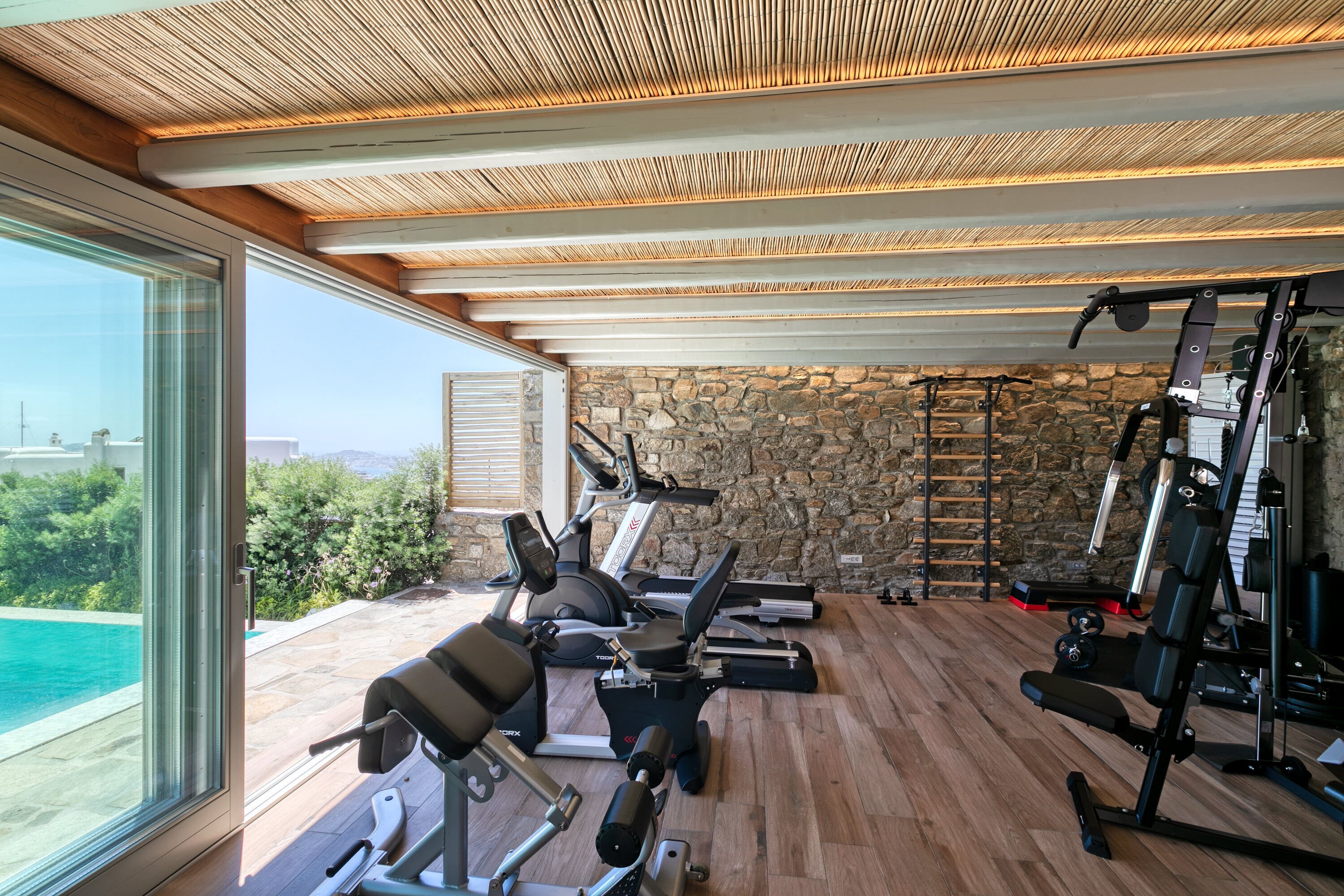 Ein Fitnessraum mit Trainingsgeräten, einer Steinwand und Blick auf ein Schwimmbad.