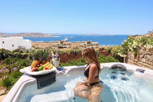 A woman relaxing in a hot tub with a fruit platter beside her, overlooking a scenic coastal view.