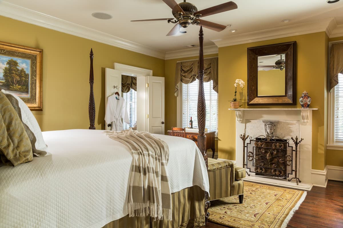 A bedroom with a large bed, a ceiling fan, a fireplace, and a mirror on the wall.