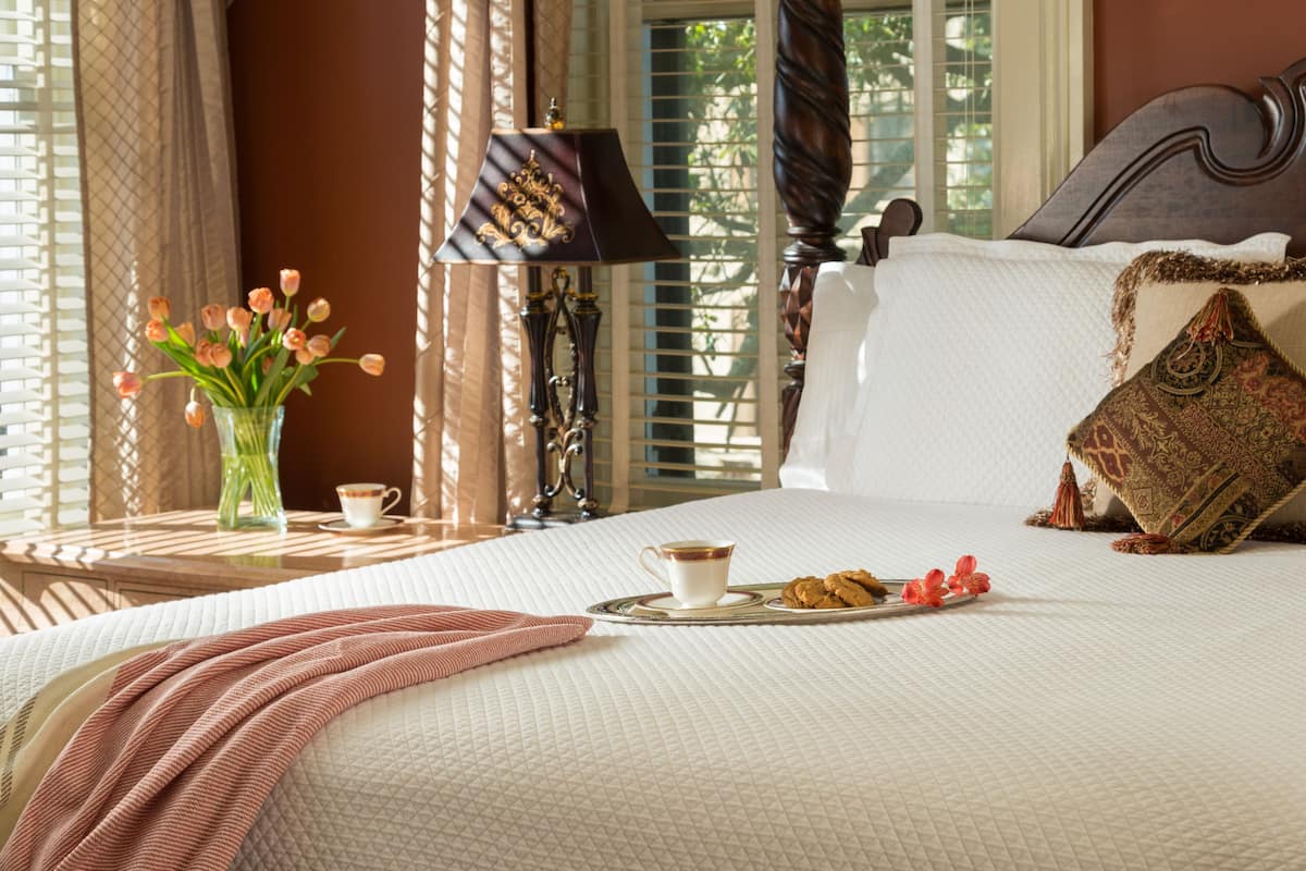 A neatly made bed with a tray of cookies and a cup of coffee, a vase of flowers, and a lamp on a bedside table.