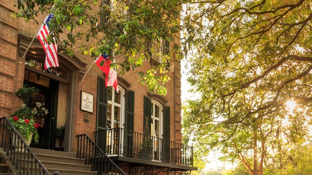 A historic building with a brick facade, a balcony with wrought iron railings, and American flags. - The Inn on West Liberty (Savannah)