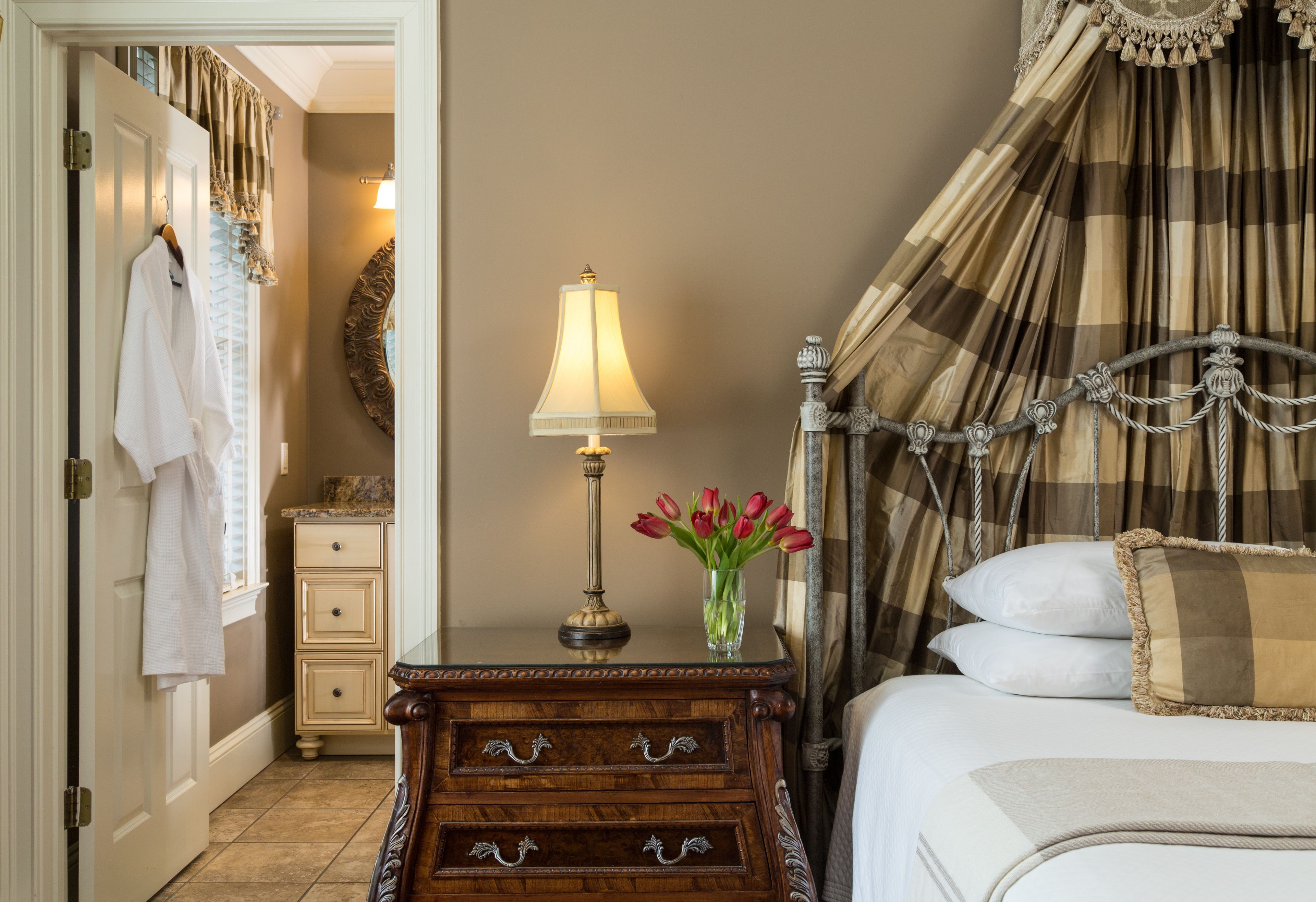 A bedroom with a bed, a nightstand with a lamp, a vase with flowers, a mirror, and a door with a towel hanging.