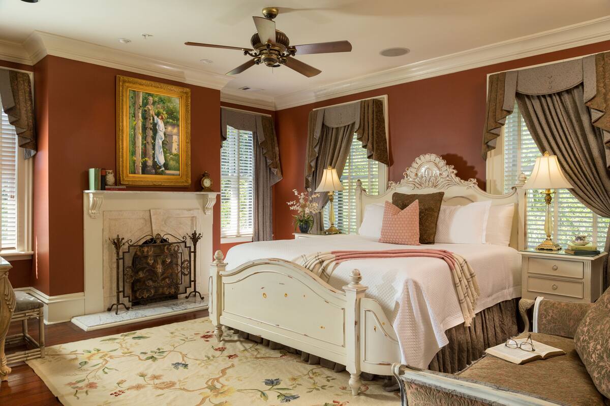 A bedroom with a white bed, a fireplace, a ceiling fan, and a large window with curtains.