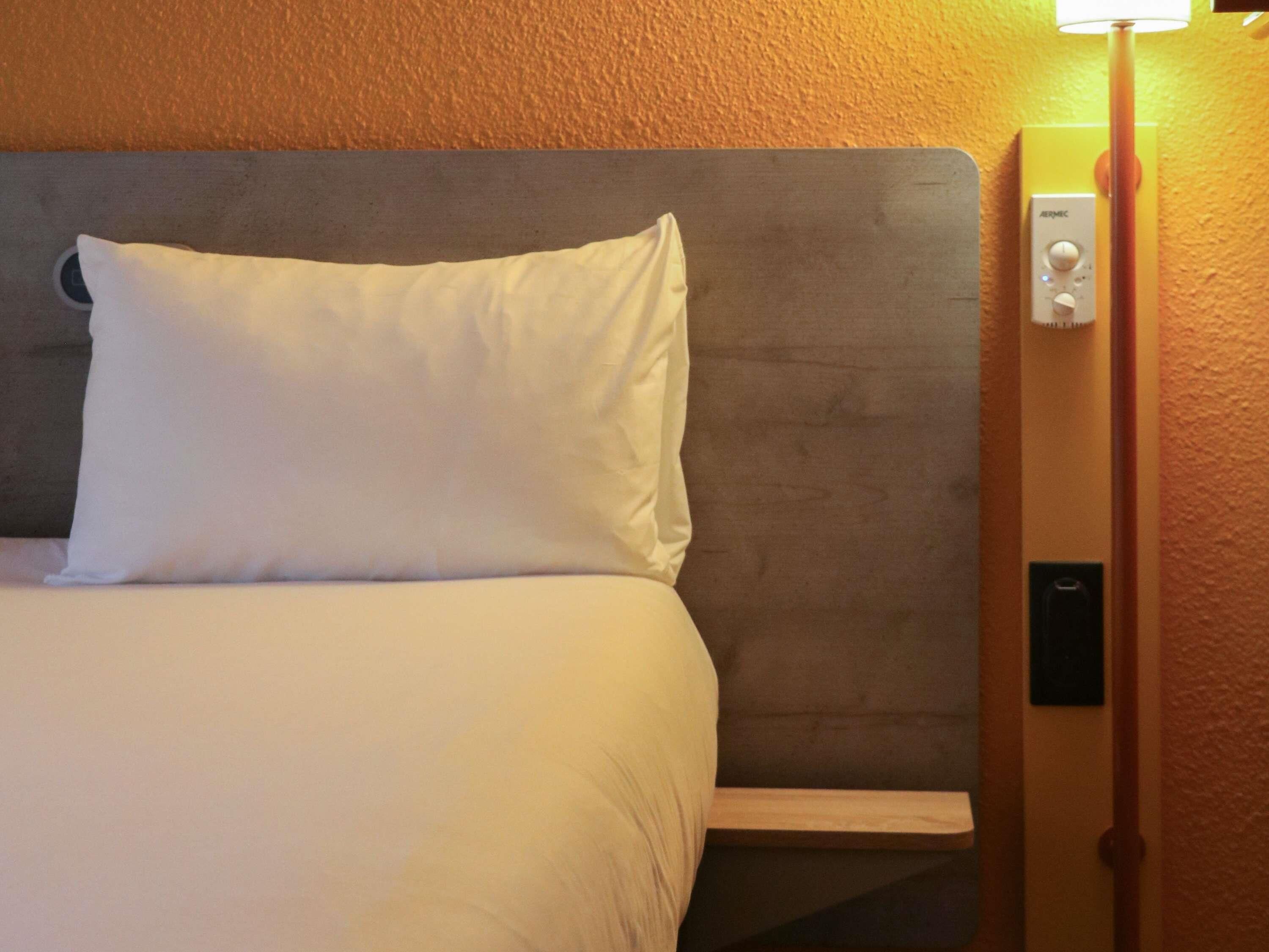 A hotel room with a bed, a nightstand, a wall-mounted thermostat, and a lamp.